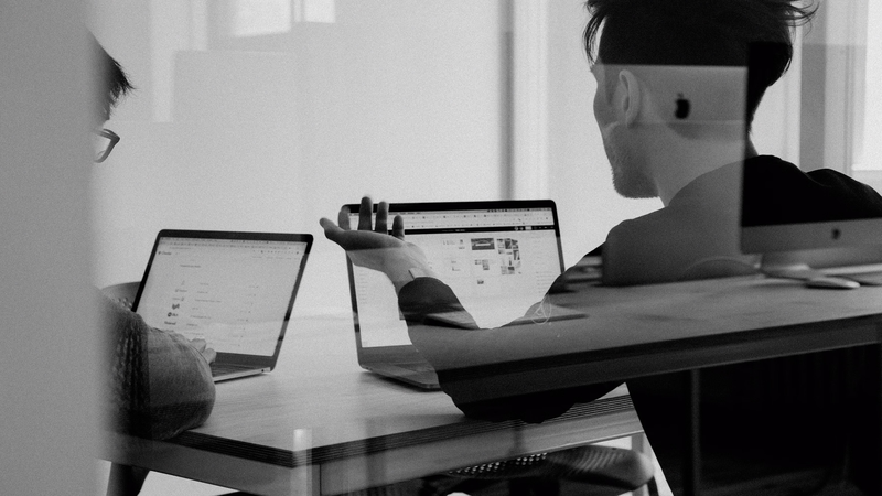 Two people discussing in front of laptops, captured through glass – symbolizing teamwork and digital collaboration.